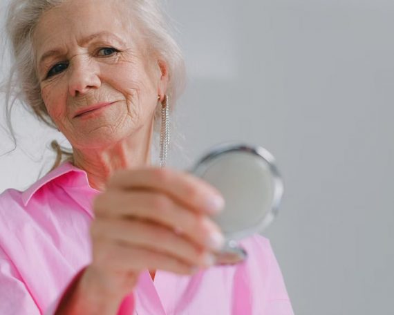 Elegant elderly woman in pink shirt holding a compact mirror indoors, reflecting joy and confidence.