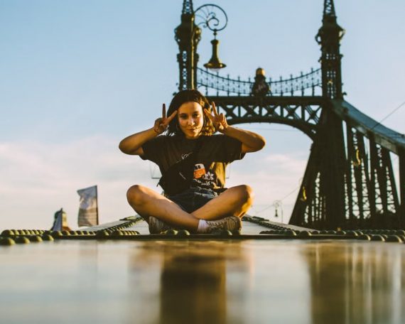 A young tourist poses with a peace sign while seated cross-legged on Budapest's Liberty Bridge during a sunny day.